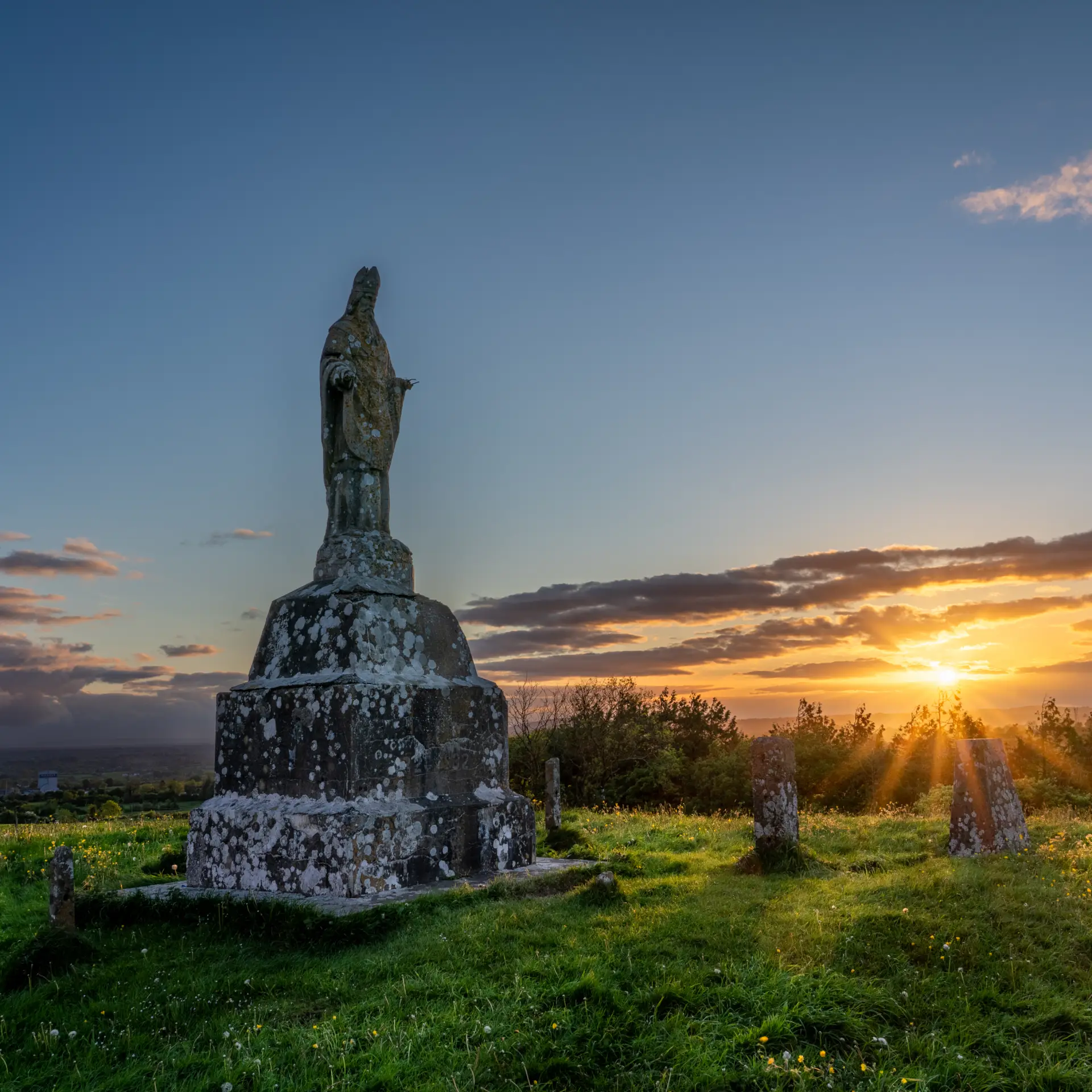 A weathered stone statue stands on a grassy hill among old gravestones, illuminated by the golden rays of a setting sun with a colourful sky and scattered clouds in the background.