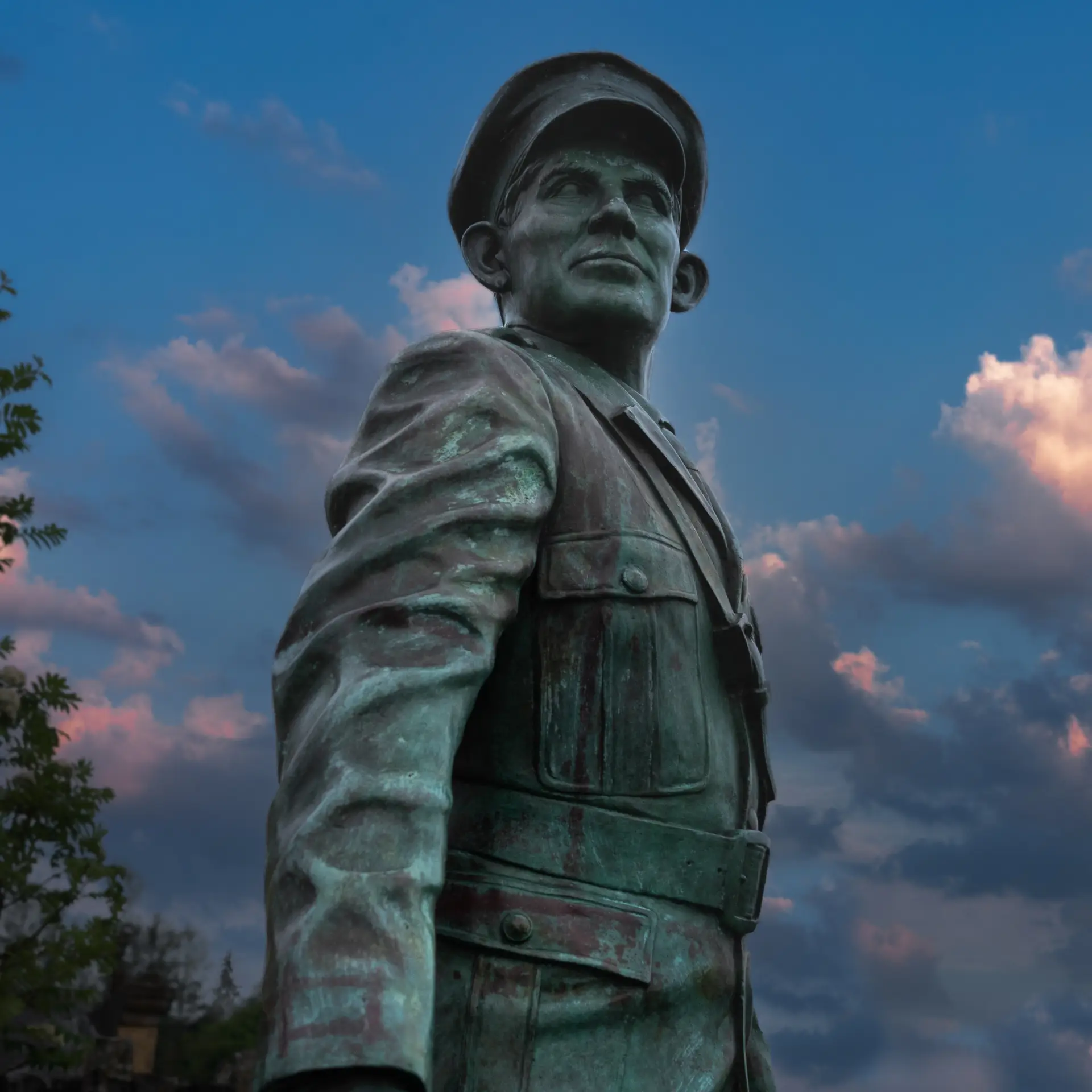 A bronze statue of a man in a military uniform and cap stands tall, looking upward with a determined expression, set against a dramatic sky with pink and blue clouds at sunset.