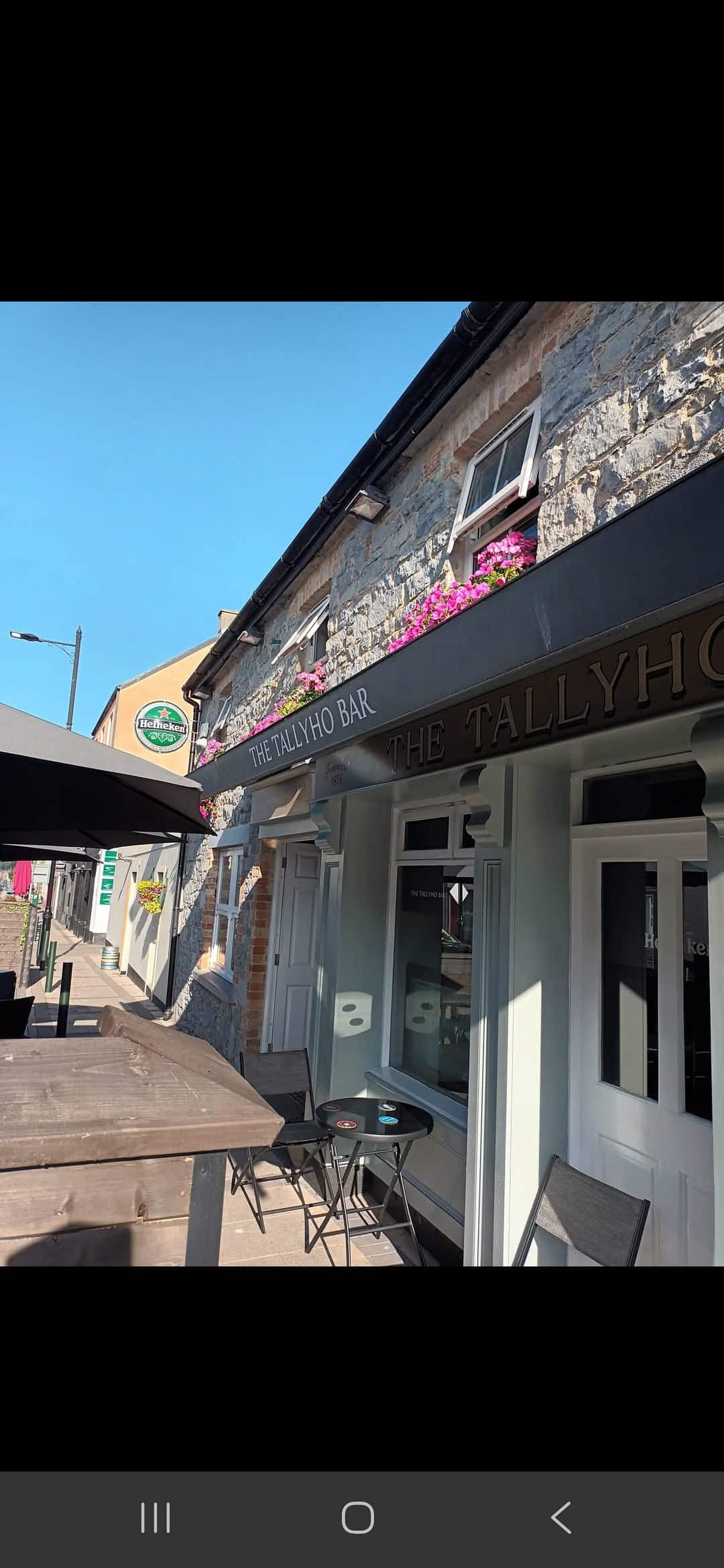 A street view of The Tallyho Bar, with outdoor seating and pink flowers in window boxes on the ground floor. The sky is clear and blue.