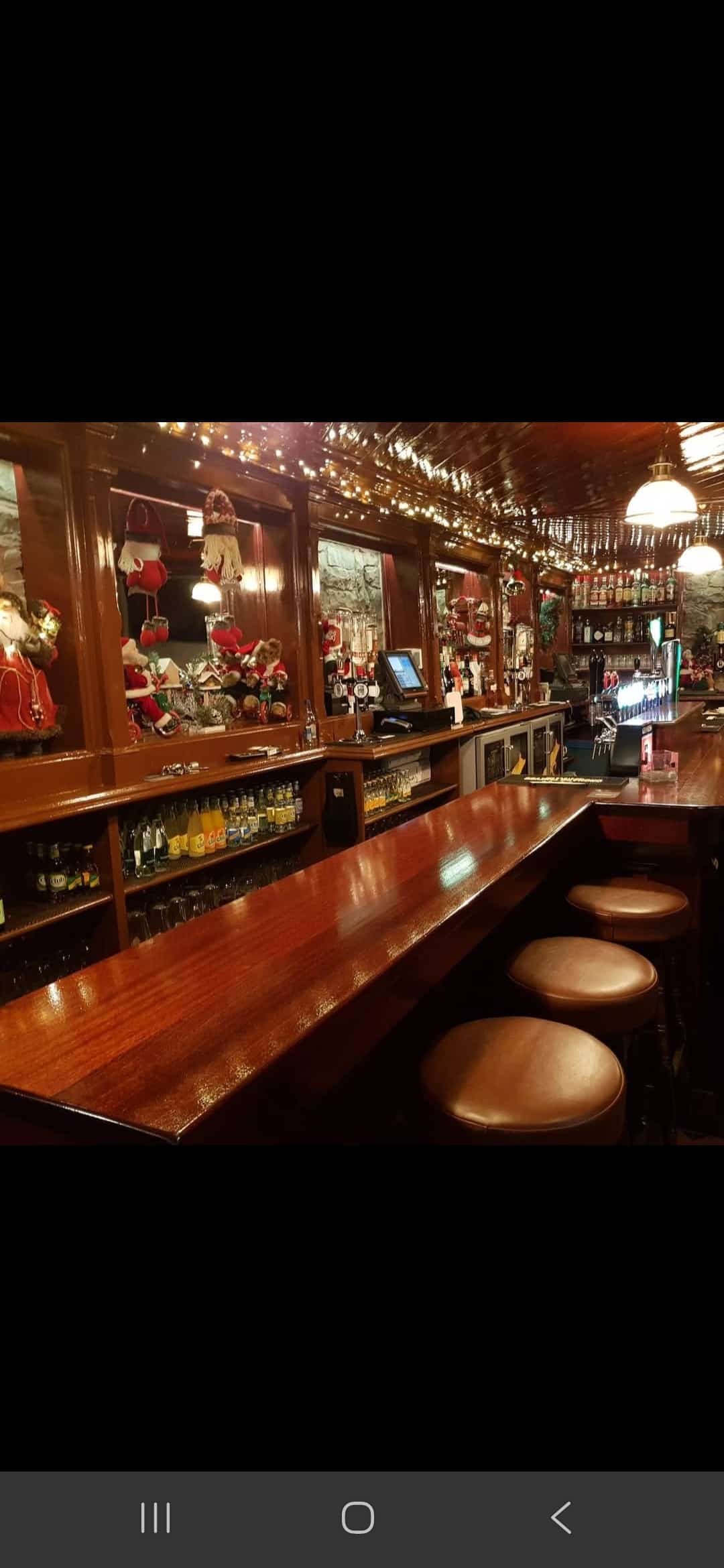 A polished wooden bar with three stools, festive Christmas decorations, fairy lights, and shelves of glasses and bottles. The bar area is warmly lit, creating a cosy atmosphere.
