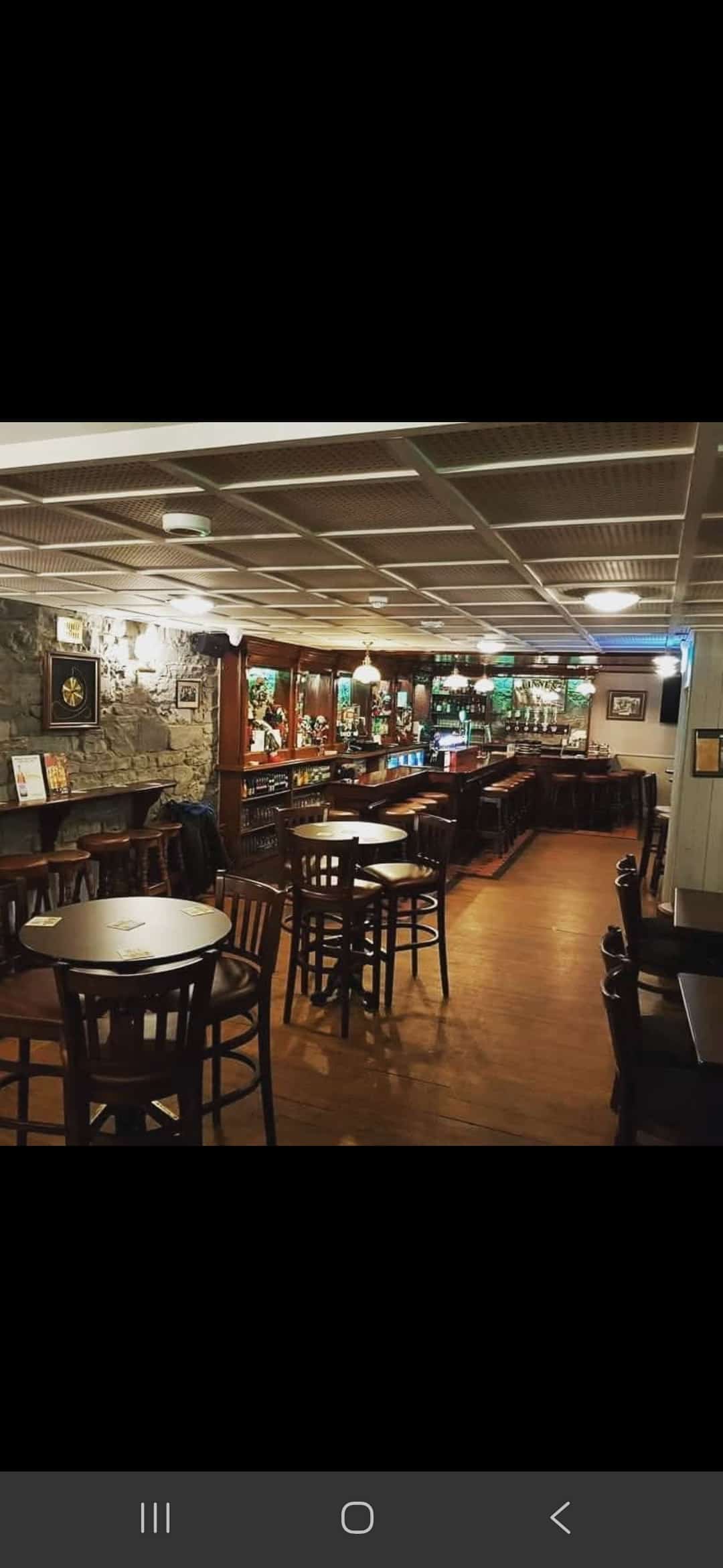 A cosy pub interior with wooden tables and chairs, stone accent walls, framed pictures, and a long bar with stools. Warm lighting creates a welcoming atmosphere.
