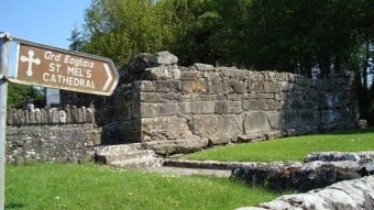 A weathered stone wall stands on grass near a wooden sign pointing to St. Mel's Cathedral, with trees in the background and sunlight illuminating the scene.