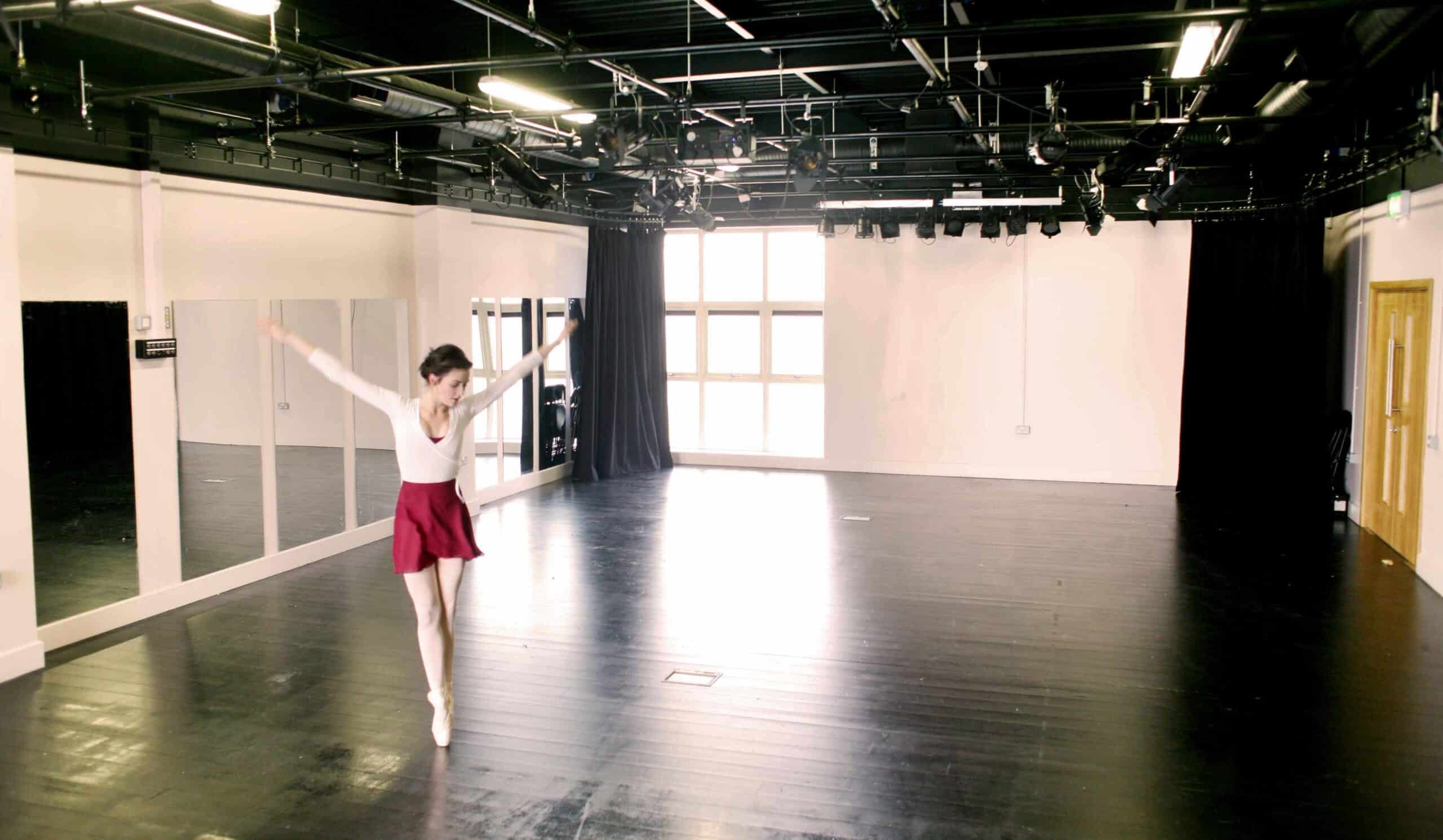 A ballet dancer in a white top and red skirt performs with arms outstretched in a spacious, empty dance studio with large windows and wooden floors.