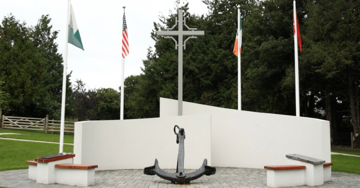 A memorial with a large anchor, three benches, a tall cross, and four flagpoles displaying different national flags, set in a landscaped park area with trees and grass in the background.