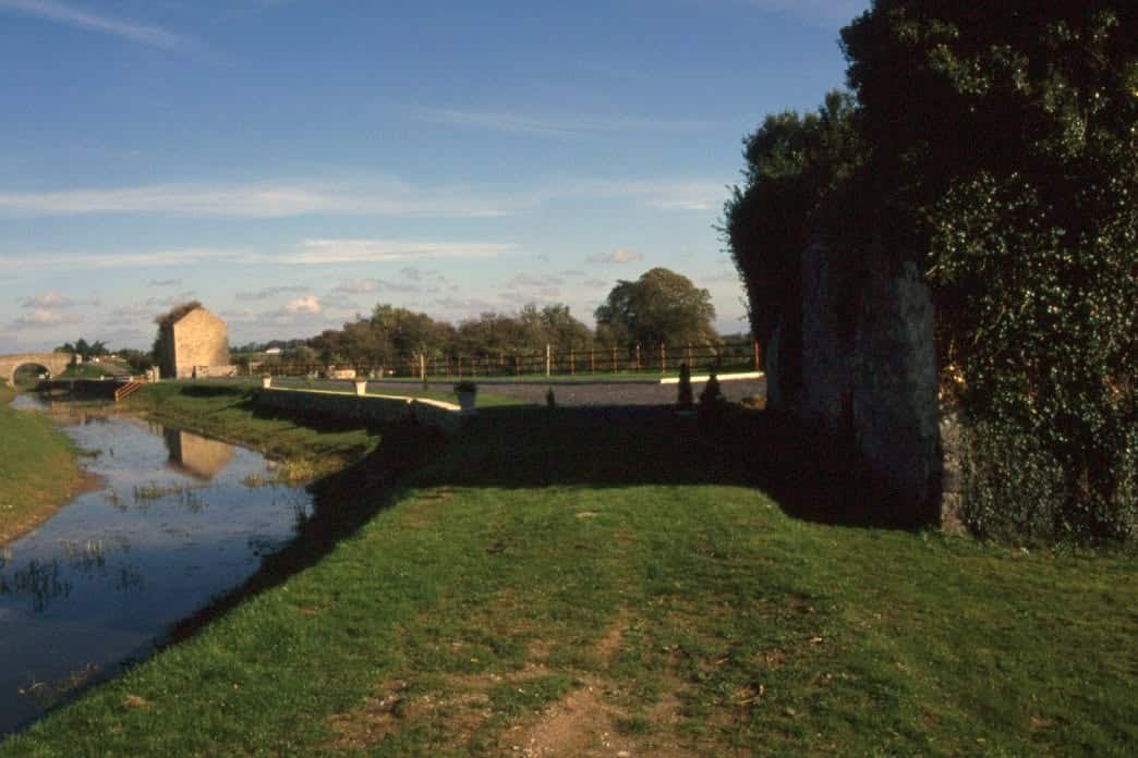 A grassy path runs beside a narrow canal with clear water. Stone buildings and trees are visible in the background to the left and right under a blue sky with scattered clouds.