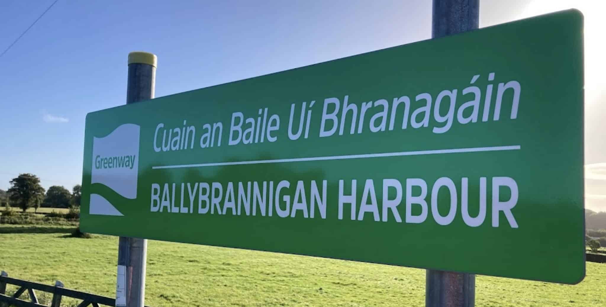 A green sign reads "Cuain an Baile Uí Bhranagáin" and "Ballybrannigan Harbour" with "Greenway" and a wave icon, placed in a grassy rural area under blue sky.