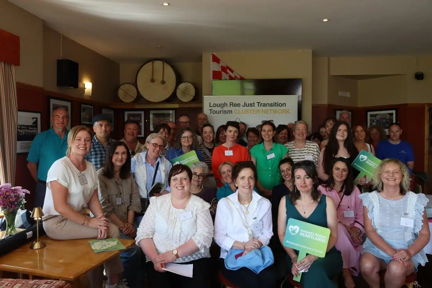 A group of people pose indoors, smiling at the camera during a tourism event highlighting Family Activities Longford. Some hold green “Hidden Heartlands” signs, and a banner reads “Lough Ree Just Transition Tourism Cluster Network.”.