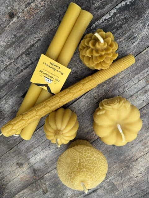 Several handmade yellow beeswax candles of various shapes, including tapers, a pine cone, a pumpkin, a rose, and a round floral design, are arranged on a rustic wooden surface.