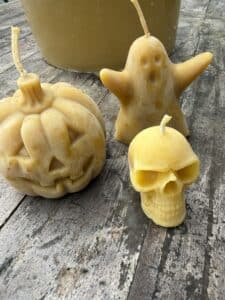 Three yellow candles shaped like a pumpkin, a ghost, and a skull sit on a weathered wooden surface.