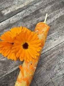A handmade beeswax candle decorated with orange petals lies on a rustic wooden surface next to two vibrant orange marigold flowers.