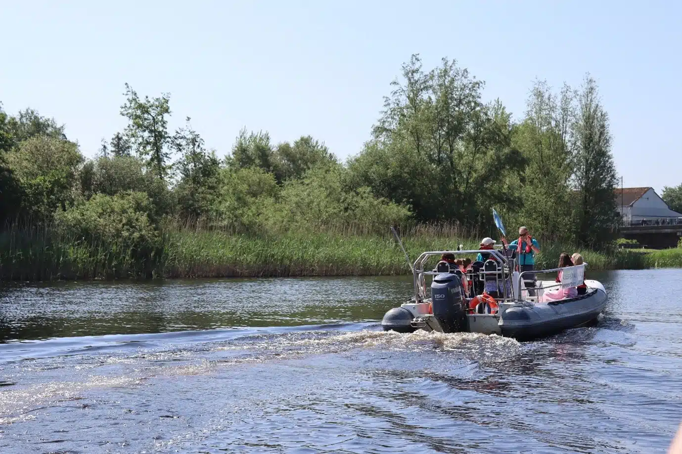 A small inflatable motorboat with several people on board moves along a calm river, leaving ripples in the water—an ideal setting for Family Activities Longford. Lush green trees and reeds line the riverbank under a clear blue sky.