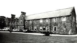 A black-and-white photo of a long, two-storey stone building with several cars parked in front and grass in the foreground.
