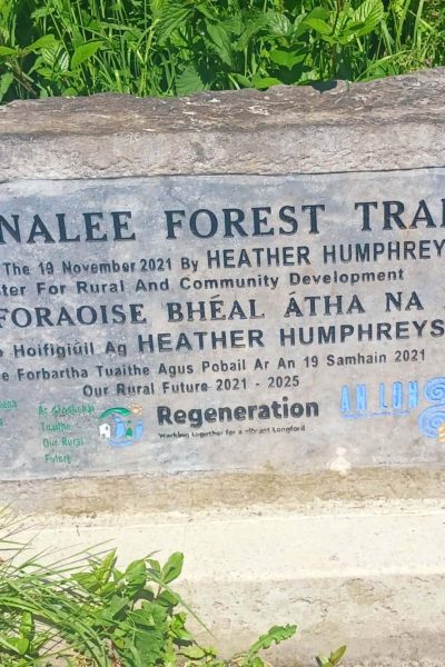 A stone sign for Ballinalee Forest Trail surrounded by grass, featuring English and Irish text about its official opening by Heather Humphreys T.D. on 19 November 2021, and various logos at the bottom.