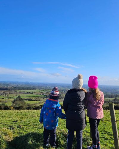 Three children in colourful winter coats and hats stand on a grassy hill, facing away from the camera and looking out over a wide, green landscape under a bright blue sky.