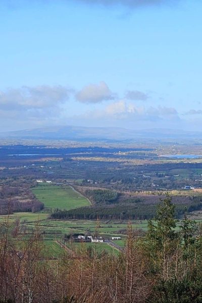 A panoramic view of a rural landscape with fields, scattered houses, and patches of woodland under a blue sky with scattered clouds; distant hills stretch across the horizon.