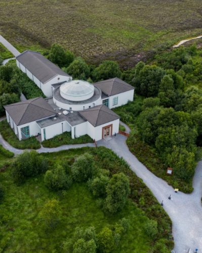 Aerial view of a white, modern building with a central glass dome surrounded by lush green trees and winding gravel paths in a rural, grassy landscape.