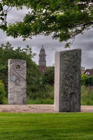 Five large carved stone pillars stand in a circle on a grassy area under a tree, with houses and a church tower visible in the background on a cloudy day.